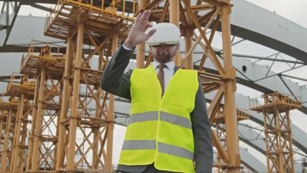Low angle shot van mannelijke bouwplaatmanager in VR-bril gebarsten tijdens het gebruik van virtual reality voor onafgewerkt gebouw met steiger - Video