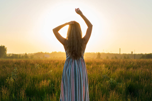 une fille dans une robe dans un champ vert dans les rayons lumineux du soleil couchant - Photo, image