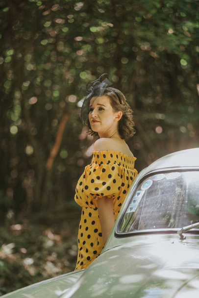 A vertical shot of a young Caucasian female performing for music video in a yellow and black dotted dress and a hairband near a retro car - Foto, afbeelding