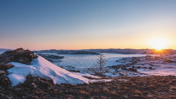 Téli hajnal Szibériában. Az ég a hegyek felett rózsaszínűvé válik. A nap sugarai ragyognak egy befagyott tó jegén. Turistabázis a parton. A földön - hó, száraz fű. Bajkál.   - Fotó, kép