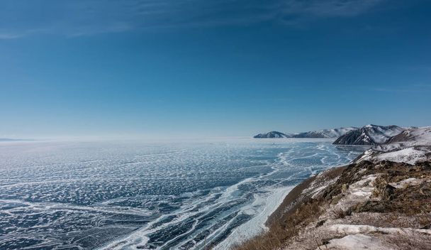 Patrones de nieve calados en el hielo azul de un lago congelado. La hierba seca crece en las colinas costeras. Cielo azul. Un día soleado de invierno. Baikal..  - Foto, imagen