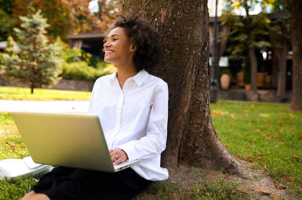 Темноволосая девушка сидит под деревом с ноутбуком - Фото, изображение