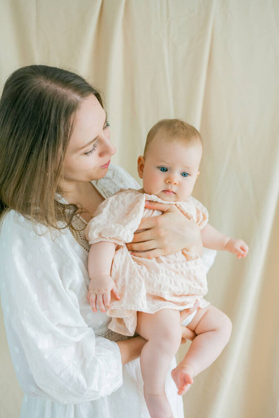 Una joven y hermosa madre con un vestido blanco sostiene a su pequeña hija de ojos azules de 5 meses en sus brazos. - Foto, Imagen