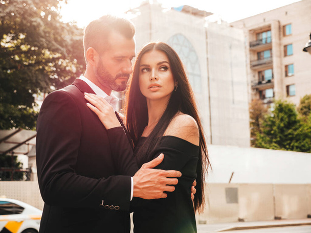 Beautiful fashion woman and her handsome elegant boyfriend in suit. Sexy brunette model in black evening dress. Fashionable couple posing in the street in Europe. Brutal man and his female outdoors - Fotografie, Obrázek