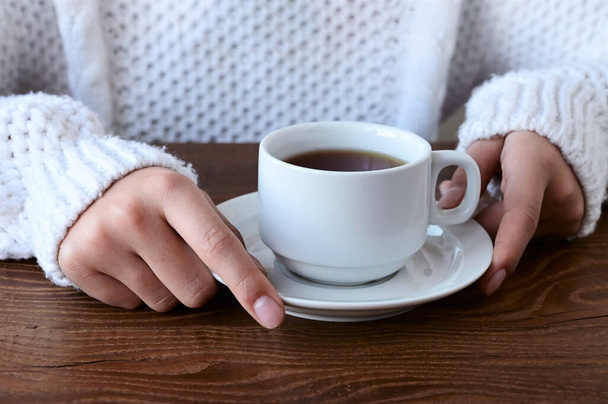 Close-up of girl's hands in warm knitted white sweater holding cup of tea on wooden table. The concept of winter comfort, relaxation - Fotoğraf, Görsel