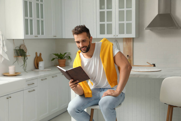 Handsome man reading book on stool in kitchen - 写真・画像