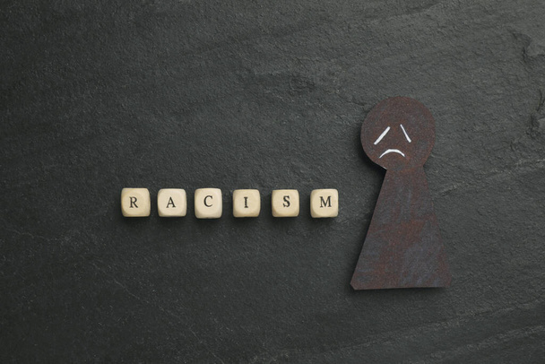 Word Racism and black paper figure with sad face on dark background, flat lay - Photo, Image