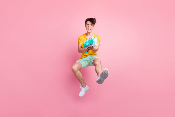 Foto van gekke gewelddadige funky man springen schot pomp waterpistool dragen geel t-shirt shorts schoenen geïsoleerd op roze achtergrond - Foto, afbeelding