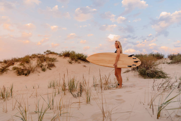 Девушка-серфер с доской для серфинга на песчаном пляже и теплыми цветами заката. Привлекательные серферши на пляже - Фото, изображение