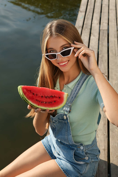 Beautiful girl with slice of watermelon on wooden pier near river - Photo, Image