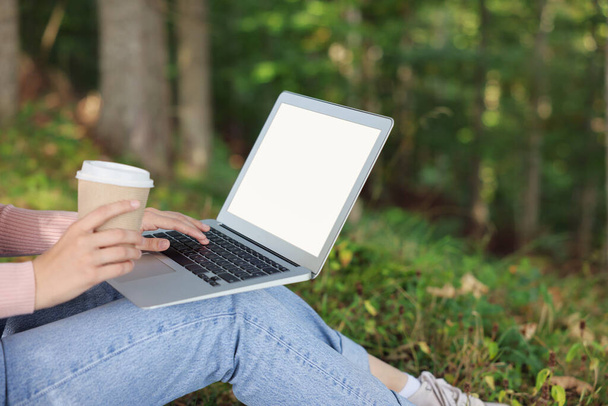 Young woman with cup of coffee working on laptop in forest, closeup - Foto, imagen