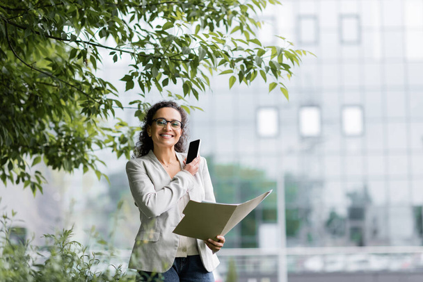 mujer de negocios afroamericana con teléfono inteligente y carpeta de papel sonriendo a la cámara bajo ramas de árboles al aire libre - Foto, Imagen