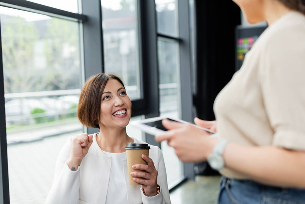 схвильована бізнес-леді тримає каву, щоб піти, показуючи жест виграшу біля розмитого афроамериканського колеги з цифровим планшетом
 - Фото, зображення