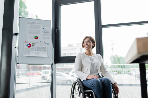 szczęśliwy businesswoman siedzi na wózku inwalidzkim w pobliżu flip chart z wykresami w biurze - Zdjęcie, obraz