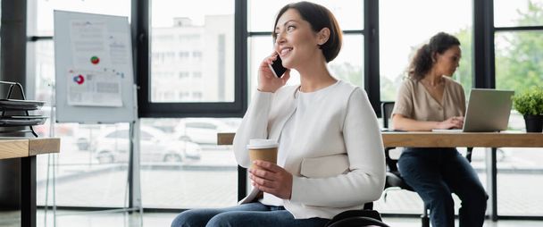mujer feliz con discapacidad sosteniendo café para ir mientras habla en el teléfono celular cerca de un colega afroamericano, pancarta - Foto, imagen