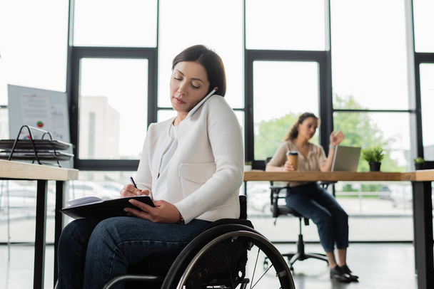 businesswoman in wheelchair writing in notebook while talking on cellphone in office - Фото, изображение