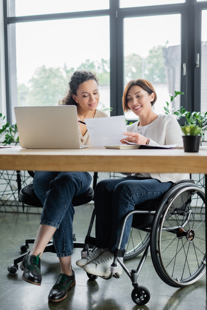 smiling woman in wheelchair working with african american colleague in office - Фото, изображение