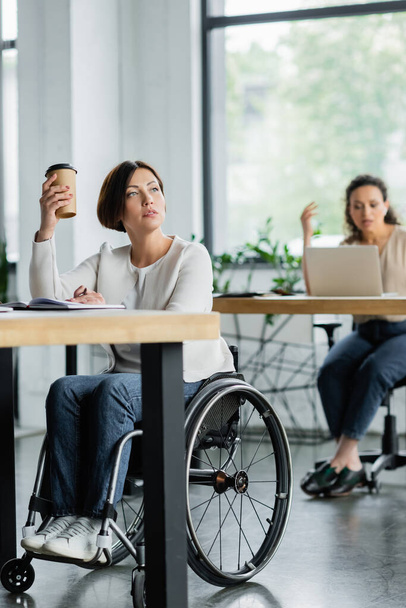 thoughtful businesswoman in wheelchair holding takeaway drink near blurred african american colleague in office - Valokuva, kuva