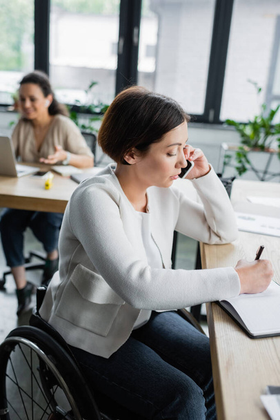 donna d'affari con disabilità fisica che scrive nel quaderno durante la conversazione sullo smartphone in ufficio - Foto, immagini