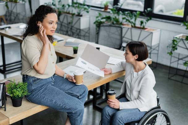 businesswoman in wheelchair looking at infographics near african american colleague sitting on desk during coffee break - Photo, Image