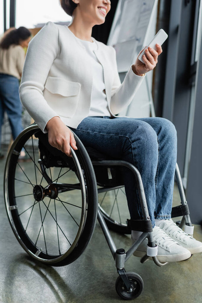 vista recortada de la mujer de negocios feliz con discapacidad física utilizando el teléfono móvil en la oficina - Foto, Imagen