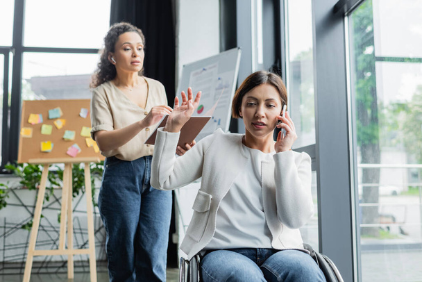 mujer de negocios en silla de ruedas hablando por teléfono móvil y mostrando gesto de espera a un colega afroamericano - Foto, imagen