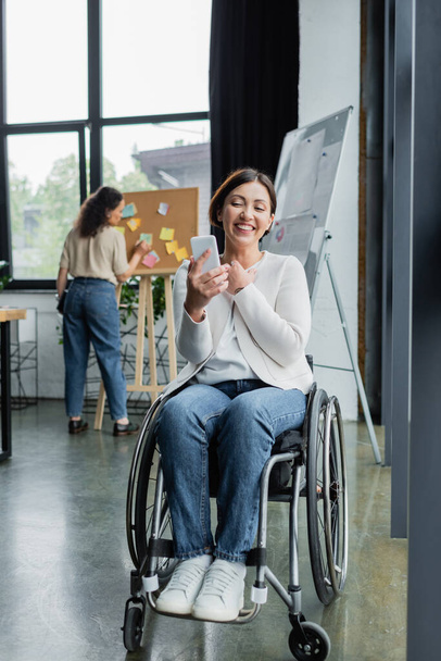 cheerful businesswoman in wheelchair looking at cellphone near african american colleague on blurred background - Photo, Image