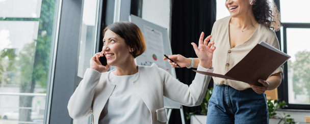 smiling businesswoman showing wait gesture to cheerful african american colleague during phone conversation, banner - Photo, Image