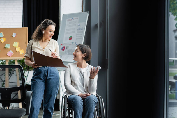 šťastný businesswoman v invalidním vozíku mluvit s africkým americkým kolegou v blízkosti rozmazané flip chart v úřadu - Fotografie, Obrázek