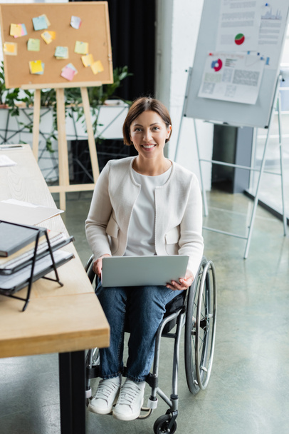 smiling businesswoman in wheelchair working on laptop near blurred corkboard and flip chart with infographics - 写真・画像