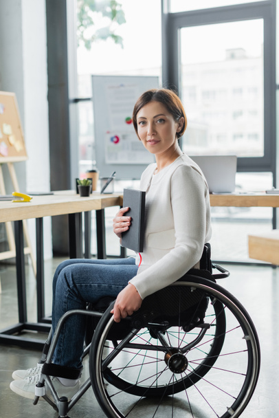 sonriente mujer de negocios con discapacidad física sosteniendo cuaderno en la oficina - Foto, imagen