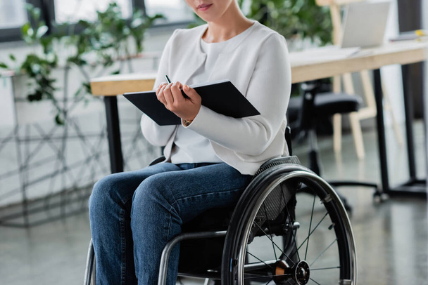 vista ritagliata di donna d'affari in sedia a rotelle scrittura in notebook in ufficio - Foto, immagini