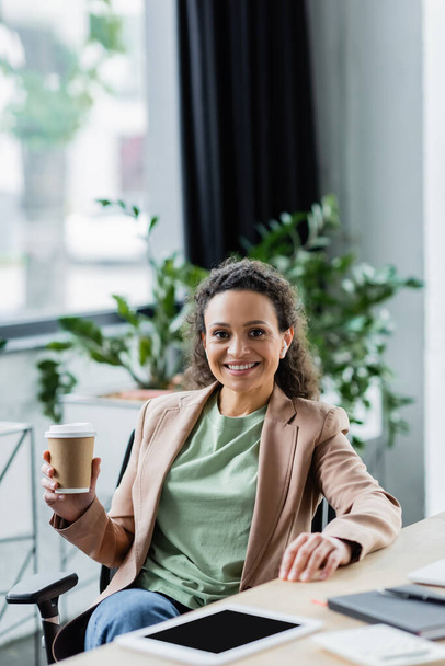 successful african american businesswoman with coffee to go smiling at camera while sitting at workplace - Fotoğraf, Görsel