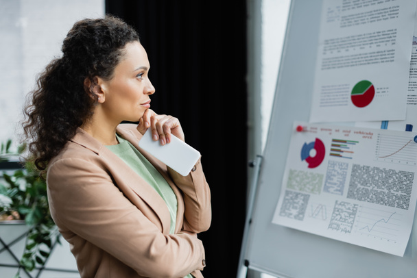 вдумчивая африканская американская бизнесвумен с мобильным телефоном смотрит на флип-чарт с аналитикой в офисе - Фото, изображение