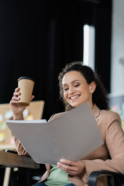cheerful african american businesswoman holding coffee to go while working with documents in office - Фото, зображення