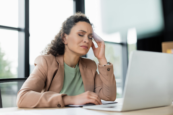 agotada mujer de negocios afroamericana que sufre de dolor de cabeza cerca de la computadora portátil borrosa en el lugar de trabajo - Foto, Imagen