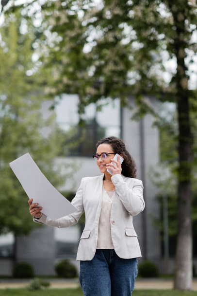mujer de negocios afroamericana positiva hablando en el teléfono inteligente mientras mira los papeles en la calle de la ciudad - Foto, imagen