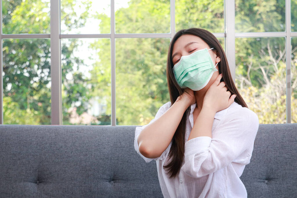 красивая азиатская женщина носить зеленую маску, чтобы предотвратить вирус сидя на диване в гостиной дома она имеет боли в шее. Концепция здравоохранения во время эпидемии коронавируса - Фото, изображение