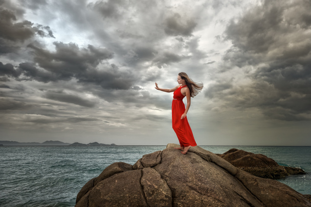 Жінка в червоній сукні стоїть на скелі з красивим видом на море
 - Фото, зображення