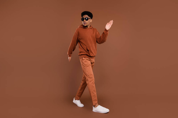 Full size profile photo of cool short hairdo young guy wave hand dress spectacles sportswear trousers sneakers isolated on brown color background - Foto, afbeelding