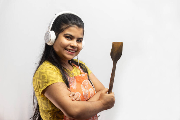 Una hermosa ama de casa india con delantal de cocina y auriculares con espátula de madera en la mano sonríe sobre fondo blanco - Foto, imagen