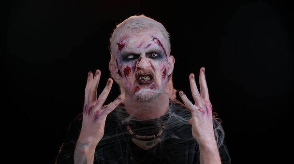 Zombie hombre en convulsiones con maquillaje con heridas falsas cicatrices en lentes de contacto blancas tratando de asustar - Foto, Imagen