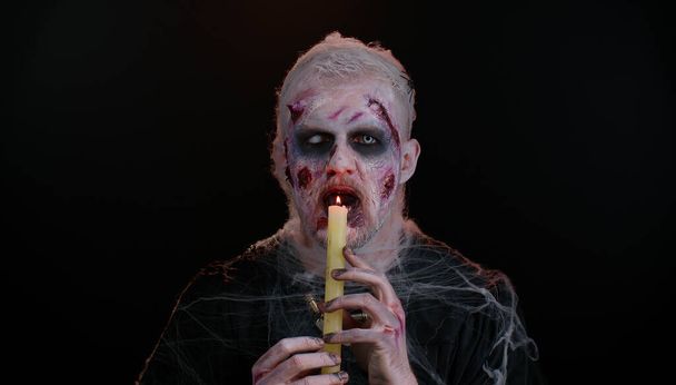 Sinistere man met vreselijke enge Halloween zombie make-up het maken van gezichten, spreuken tovert over een kaars - Foto, afbeelding