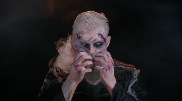 Sinister man with horrible scary Halloween zombie makeup in costume blows smoke from nose and mouth - Photo, Image