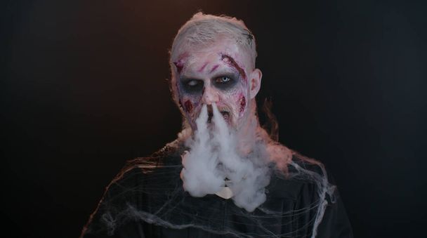 Τρομακτικός τραυματισμένος αποκριάτικος απέθαντος άντρας φυσάει καπνό από τη μύτη και το στόμα, χαμογελάει τρομερά - Φωτογραφία, εικόνα