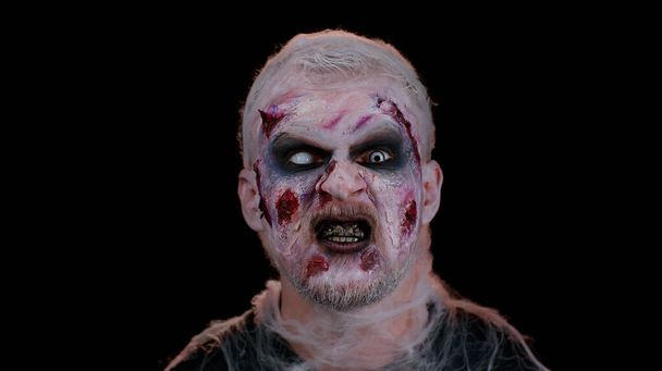 Zombie hombre con cicatrices heridas y lentes de contacto mirando la cámara hace clic en sus dientes tratando de asustar - Foto, imagen