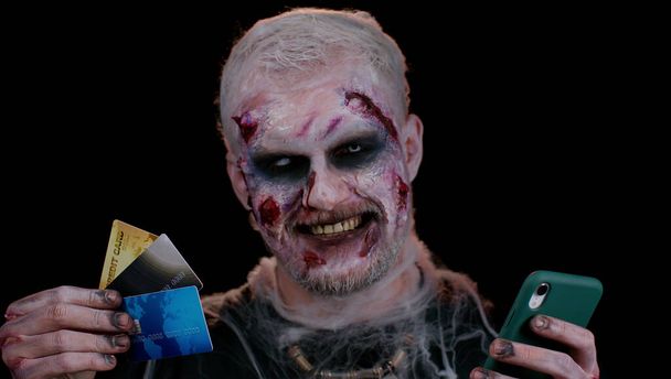 Sinister muž Halloween zombie pomocí kreditní bankovní karty a smartphone při nákupu on-line nakupování - Fotografie, Obrázek