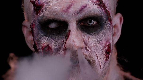 Zombie hombre con maquillaje con cicatrices heridas y lentes de contacto blancos sopla humo de la boca, sonriendo - Foto, imagen