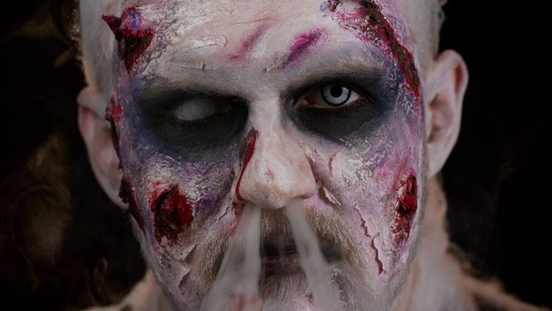 Зомби человек с макияжем со шрамами от ран и белые контактные линзы дует дым изо рта, улыбаясь - Фото, изображение