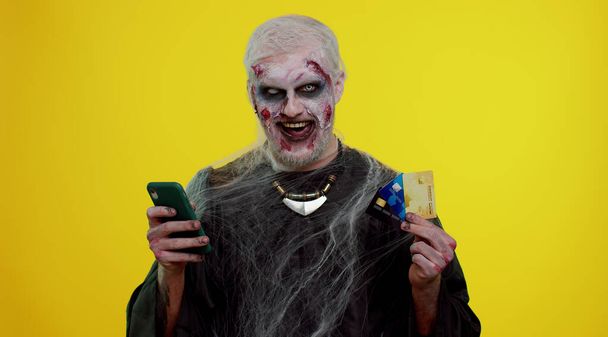 Sinister man Halloween zombie using credit bank cards and smartphone while purchases online shopping - Photo, Image